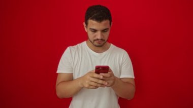Zaferi kutlayan mutlu İspanyol genç adam, kazanan bir gülümseme ve izole edilmiş kırmızı arka plan üzerinde akıllı telefon kullanarak el kaldırdı.