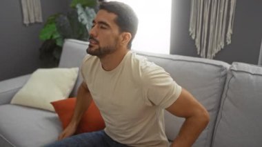 Sakallı genç adam, modern oturma odasındaki gri koltukta rahatsız hissediyor.