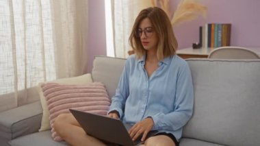 Kadın bilgisayarını kapatıp kameraya gülümsüyor ve modern oturma odasında kanepede oturuyor.