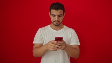 Genç İspanyol asıllı adam akıllı telefonuyla stresli ve hayal kırıklığına uğramış, el üstünde öfke ve şaşkınlık içinde izole kırmızı arka planda.