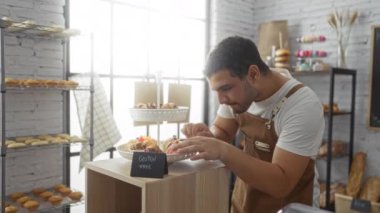 Genç adam rahat bir fırında gülümseyip kollarını kavuşturuyor. Glütensiz hamur işleri sergileniyor ve arka planda ekmek rafları var.