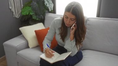 Oturma odasındaki bir kanepede oturup telefonla konuşan bir kadın not defterine yazı yazıyor..