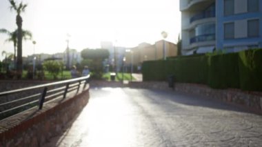 Açık hava parkı sahnesinde iki bulanık insan yeşil çitlerle çevrili bir yolda yürüyor ve huzurlu bir ortamda parlak güneş ışığı altında konutlar inşa ediliyor.