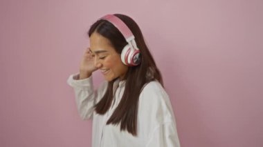 Pembe kulaklık takan genç bir kadın gülümsüyor ve açık beyaz bir gömlek içinde pembe bir arka plana karşı müzikten hoşlanıyor..