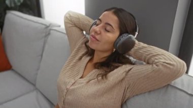 Kanepede oturmuş kulaklıklarıyla müzik dinleyen rahat oturma odasında, huzurlu anın tadını çıkaran bir kadın..