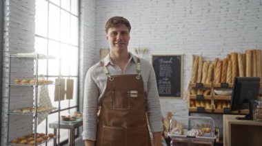 Önlüklü genç adam fırının içinde gülümsüyor ve arka planda ekmek ve hamur işleri var.