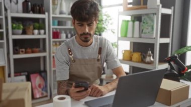Delikanlı, bir ev dekorasyonu dükkanında masa başında telefon ve dizüstü bilgisayar kullanıyor.