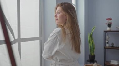 Kaplıca sağlık merkezinde beyaz bornoz giyen bir kadın. Pencereden dışarı bakarken iç mekan bitkileriyle birlikte. Huzurlu ve rahatlatıcı bir atmosfer izlenimi veriyor..