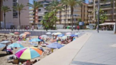 Yazın kumsala gidenler, açık hava kentsel ortamında renkli şemsiyeler altında güneşli bir günün tadını çıkarıyorlar. Arka planda palmiye ağaçları ve konut binaları var.