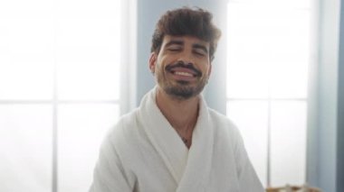 Beyaz bir bornoz ve küpe takmış, kaplıcaya doğru gülümseyen sakallı genç İspanyol bir adam.