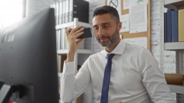 Ofiste akıllı telefondan konuşan, odaklanmış ve profesyonel görünen bir adam, bir iş konuşmasına dahil olurken, raflar, kağıtlar ve bir bilgisayar ile çevrili, yoğun bir iş yerini tasvir ediyor..