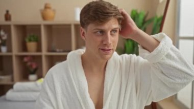 Spa Sağlık Merkezi 'nde bornozlu genç bir adam saçlarını rahat bir ifadeyle, bitki ve dekorla iç mekanda düzeltiyor.