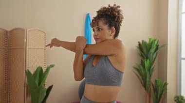 Bir kadın sağlık merkezinde bitkilerle esniyor, kapalı alanlarda egzersiz yapıyor, sağlık ve spor kıyafetleri giyip rahat bir yüz ifadesi sergiliyor..