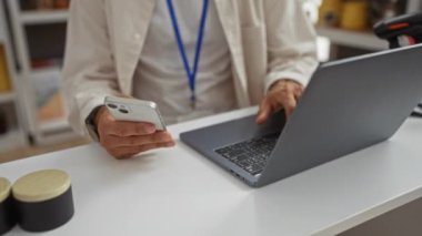 Sakallı genç adam, dekoratif eşyalarla dolu bir mağazada akıllı telefon ve dizüstü bilgisayar kullanıyor.