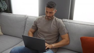 Rahat bir oturma odasında genç bir adam dizüstü bilgisayarı kapatıyor, rahat bir ifadeyle kanepeye yaslanıyor, evde huzurlu bir an sergiliyor..