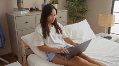 Yatak odasında dizüstü bilgisayar kullanan gözlüklü genç bir kadın ev ortamında rahat şort ve beyaz tişört giyerek laptopu kapatarak uyumaya hazırlanıyor..