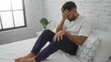 Genç adam, modern bir yatak odasında beyaz tuğla duvarlı ve yanında bir bitki olan bir yatakta otururken endişeli görünüyor..