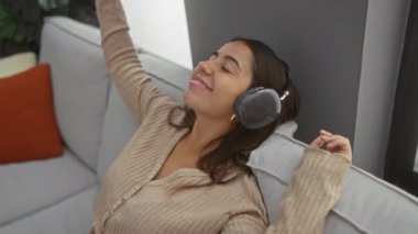 Oturma odasındaki bir koltukta kulaklıkla müzik dinleyen bir kadın, rahat bir evde keyif ve rahatlık ifade ediyor..