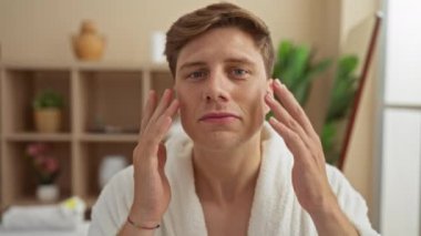 Genç bir adam, sağlık merkezinde beyaz bornoz giyip, yüz kremi sürüyor ve masaj yapıyor.