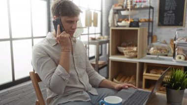 Akıllı telefondan konuşan ve arka planda çeşitli ekmek ve hamur işleri sergilenen bir fırın ortamında dizüstü bilgisayar kullanan genç adam.
