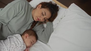 Bir kadın, uyuyan bir bebekle yatakta uzanıyor, rahat bir yatak odası ortamında huzurlu bir aile anını yakalıyor, sevgiyi ve şefkati ön plana çıkarıyor..