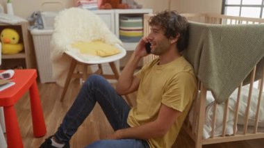 Genç adam şık bir yatak odasında oturmuş bebek beşiğinin yanında telefonla konuşuyor renkli oyuncaklar ve modern mobilyalar dahil, sıcak ve rahat bir aile ortamı yaratıyor..