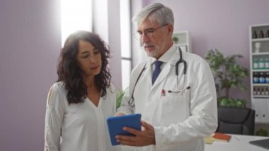 Beyaz önlüklü doktor, modern klinikteki bir kadına dijital tablet gösteriyor. Steteskopu içeride görünüyor. Sağlık hizmetleri etkileşimini vurguluyor..