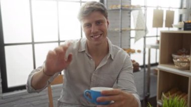 Genç adam gülümsüyor ve elinde mavi bir kupayla arka planda fırında pişmiş yiyecekler sergiliyor.