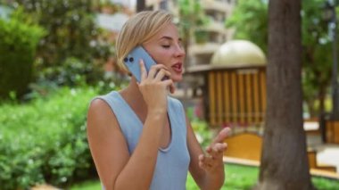 Arka planda ağaçlar ve binalar olan bir şehir parkında cep telefonuyla konuşan genç bir kadın.