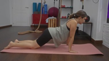 Spor salonundaki pembe bir paspasın üzerinde yoga yapan genç bir kadın. Arka planda çeşitli spor aletleri var.