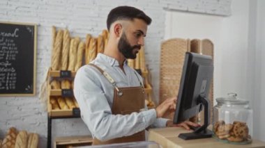 Fırın tezgahında çalışan yakışıklı İspanyol bir adam. Arka planda ekmek ve kurabiyeler var. Bilgisayar kullanıyor.