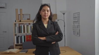 Ofiste kendine güvenen, siyah ceket giyen, kollarını kavuşturup gülümseyen, arka planda kitaplığı, masası ve lambası olan, profesyonel ve yaklaşılabilir tavırları olan bir kadın.