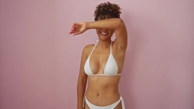 Genç İspanyol kadın bikini giyiyor ve neşeli bir şekilde pembe arka planda dikiliyor, neşeli bir yüzle gülümsüyor ve neşeli bir ifade sergiliyor.