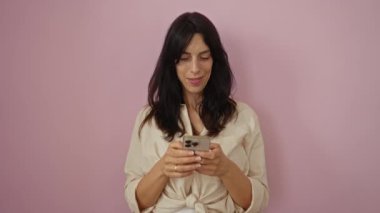 Pembe arka plan üzerinde akıllı telefon kullanan bir kadın, sürpriz ve normal bir jest de dahil olmak üzere çeşitli tepkiler gösterir, günlük bej gömlek giyer, yirmili yaşların ortasında, kapalı alanda izole edilmiş