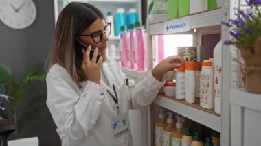 Eczanede çalışan, telefonla konuşan, raflarda ürünleri düzenleyen laboratuvar önlüğü ve gözlük takan bir kadın..