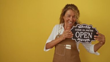 Orta yaşlı bir kadın, elinde açık tabela tutarken gülümsüyor sarı arka plan, kahverengi önlük takıyor.