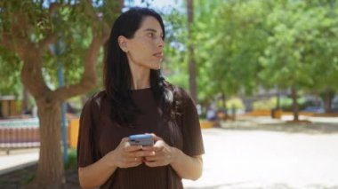 Güneşli bir günde arka planda ağaçlarla parkta akıllı telefon kullanan bir kadın.