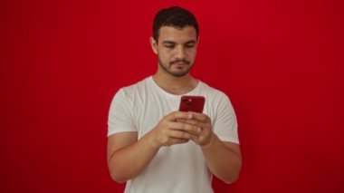Genç İspanyol adam akıllı telefonuyla işaret ediyor, mutlu ve gururlu, izole edilmiş kırmızı arka planda gülümsüyor.