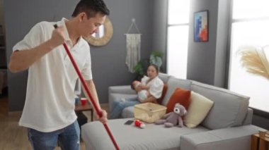 Kadın, modern oturma odasında bebek emzirirken erkek temizlik yapıyor. Aile dinamiklerini rahat ve evcil bir ortamda sergiliyor. Lüks oyuncaklarla sade bir dokunuş ekliyor..