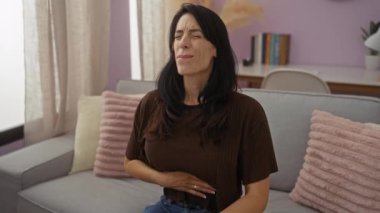 Oturma odasındaki kanepede karın ağrısı çeken bir kadın. Perdeleri ve yastıkları var. Acısını ya da hastalığını ifade ediyor.