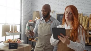 Erkek ve kadın birlikte bir fırında çalışıyor, tablet üzerinde tartışıyorlar, arka planda ekmek ve hamur işleri, önlük giyiyorlar ve modern bir dükkanın içinde duruyorlar.