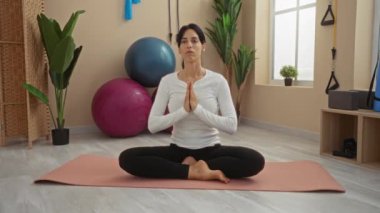 Spor salonunda meditasyon yapan bir kadın yoga minderinde gözleri kapalı ve elleri dua pozisyonunda otururken fitness malzemeleriyle çevrili ve huzurlu sağlık pratiği için doğal ışık kullanıyor.