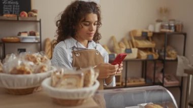 Fırın odasında çeşitli ekmek ürünleriyle çevrili kırmızı bir telefon kullanan kahverengi önlüklü ve kıvırcık esmer saçlı genç bir kadın..