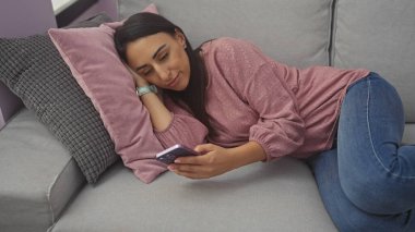 Rahat bir kadın evdeki koltukta uzanıyor, akıllı telefonunu kullanıyor, rahatlık ve modern yaşam tarzını somutlaştırıyor..