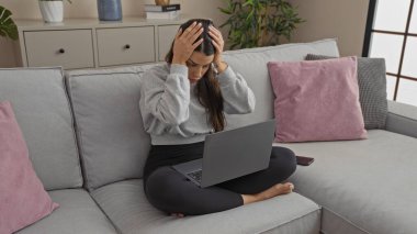 Genç bir İspanyol kadın oturma odasındaki kanepede oturur, dizüstü bilgisayarına stresli bir ifadeyle odaklanır, etrafı ev bitkileriyle ve dekorla çevrilidir..