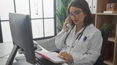 Hastane kliniğinde, gözlüklü ve beyaz önlüklü genç bir İspanyol kadın doktor masasında dokümanları okurken telefonla konuşuyor..