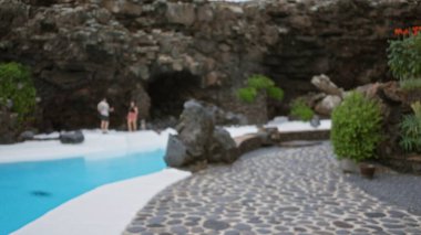 Lanzarote 'daki açık hava sahnesinde volkanik bir mağaranın etrafında yeşillik ve havuz kenarı olan kayalık bir zemin üzerinde bulanık bir adam ve kadın görülüyor.