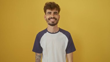 Sakallı, gülümseyen, genç bir İspanyol adam sarı bir arka plana karşı duruyor, sıradan ve dostane bir hava yayıyor..