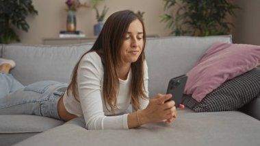 Genç, çekici bir kadın oturma odasındaki bir kanepede uzanmış, akıllı telefonuna odaklanmış, rahat ve samimi bir sahne yaratıyor..