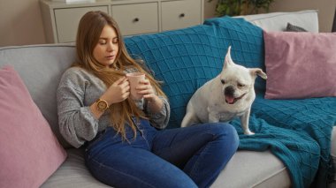 Sarışın, beyaz bir kadın oturma odasındaki kanepede bir fincan kahveyle rahatlıyor. Yanında köpeğiyle birlikte evinde rahat bir ortam yaratıyor..